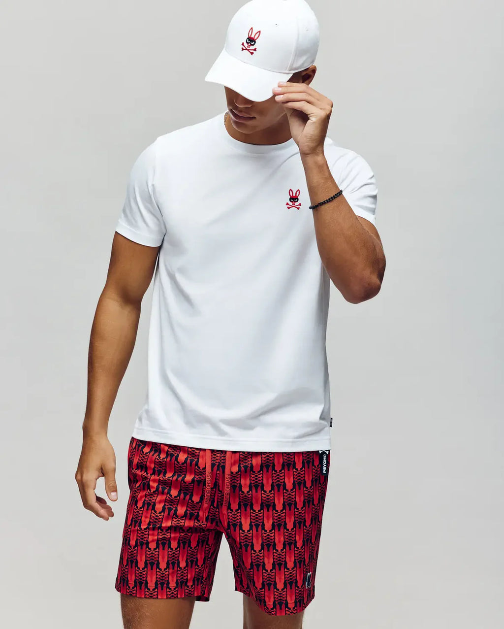 Person wearing a white t-shirt, red patterned shorts, and a white cap on a plain background
